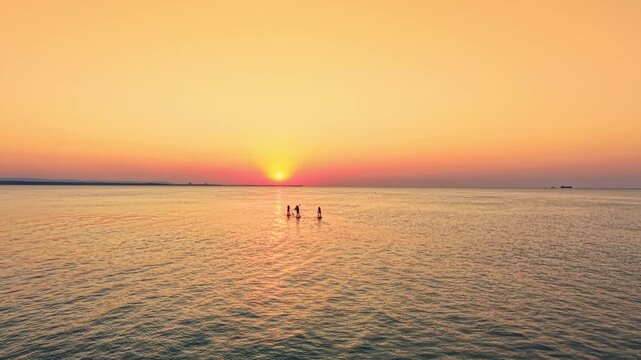 Groups of people stand in the water as the sun sets over the horizon. The sky shows colors of orange and pink while the waves gently splash around them.