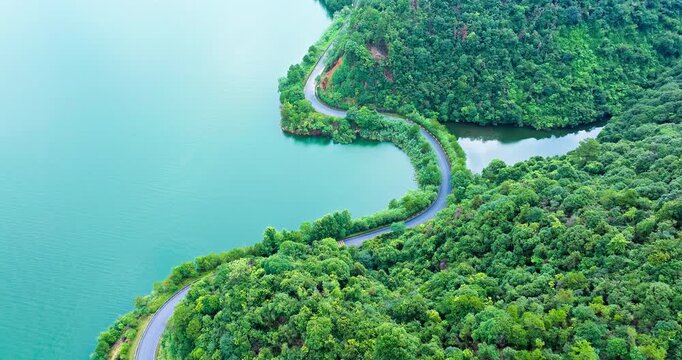 Aerial view of a winding paved road beside a calm turquoise lake, bordered by dense forested hills in bright sunlight.