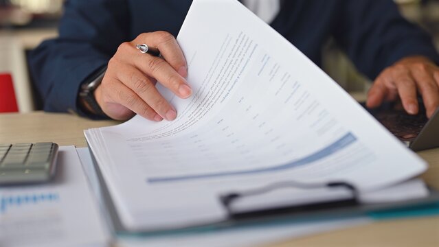Executive handles analysis reviewing financial document in office, doing corporate data analysis examining paper document, holding pen near laptop, completing risk analysis, preparing tax audit.
