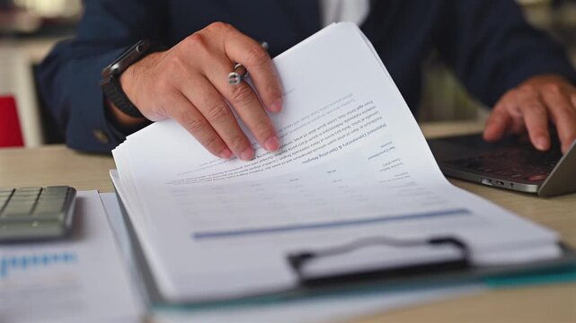 Executive performs audit procedures examining financial report sheets, managing audit tasks beside laptop, reviewing tax report details inside finance audit office.