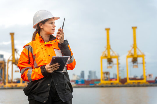 Female port engineer using walkie talkie and digital tablet managing operations at shipping container terminal, logistics manager in safety gear communicating via radio at harbor site