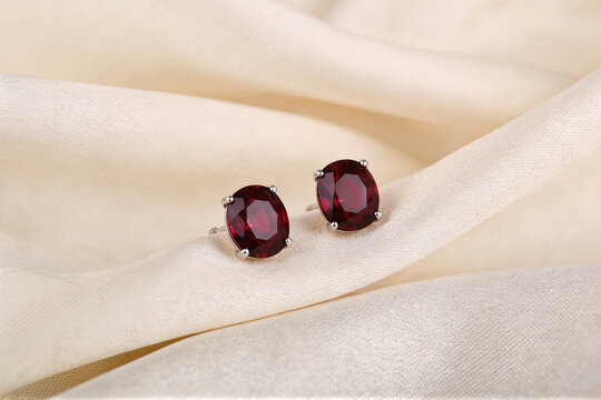 Pair of oval-cut red garnet stud earrings in silver prong settings on cream fabric, vivid jewelry close up
