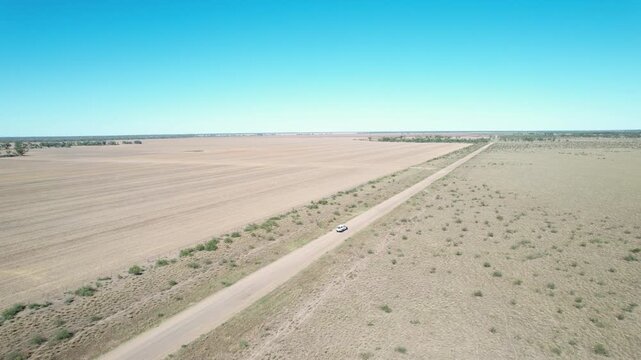 Aerial view of a lone white vehicle driving along a long remote dirt road in the dry Australian outback landscape under clear blue skies.