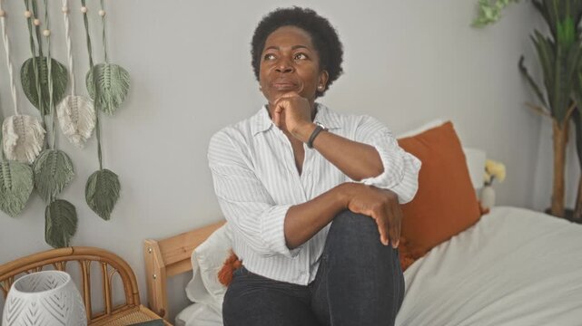 Woman sitting on bed with hand to chin in building decorated with macrame and plants; calm reflection.