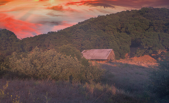 A rustic barn sits peacefully at the edge of a lush green hill under a dramatic sunset sky. The vibrant orange and pink hues illuminate the landscape