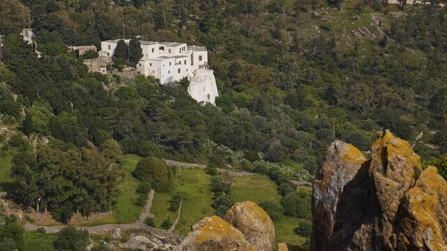 White building from a distance in a wooded landscape with rocks in the foreground, Monastery of the Revelation, Moni tis Apokalipsis, World Heritage Site 1999, Patmos, Dodecanese, Greek Islands, Greece