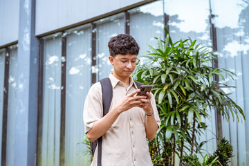 A young man with curly hair stands outside, focused on his smartphone, surrounded by. © Firman Maulana Ihsan