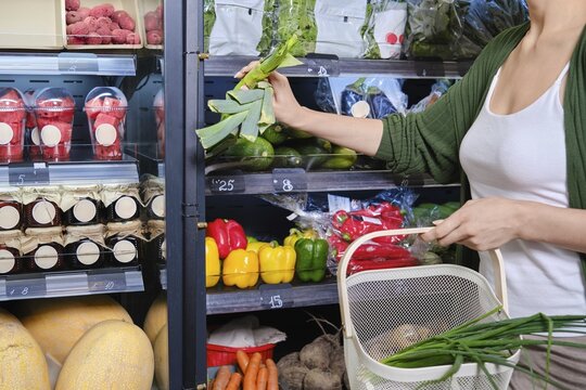 Person picks fresh vegetables from shelf in grocery store. Other items include fruits and packaged goods. Shopping basket is nearby. Store has bright lights and organized displays
