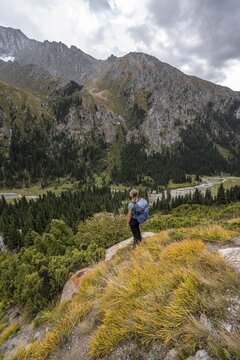 A female hiker stands on a grassy slope against an impressive mountain backdrop under a cloudy sky, Chong Kyzyl Suu Valley, Terskey Ala Too, Tien-Shan Mountains, Kyrgyzstan