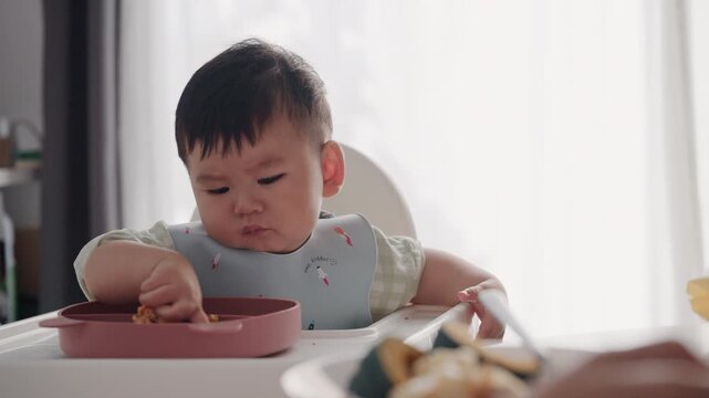 Asian Baby Using Finger to Feed Herself a Food with Baby Led Weaning Concept.Cute Asian baby boy eating finger food in high chair.