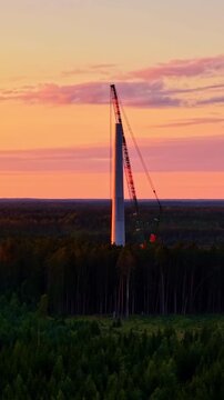 A wind turbine under construction in forest area on a sunset background.