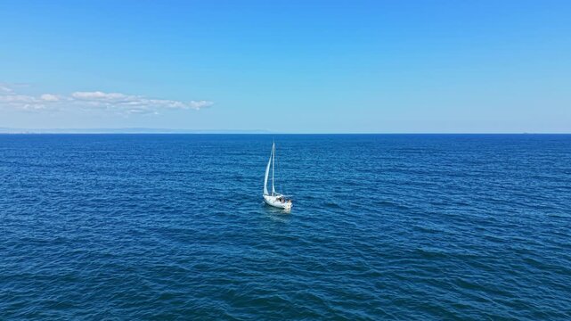 A sailboat moves across a vast ocean under a clear blue sky. The sun shines bright, and waves create a smooth surface. The horizon stretches far in the distance.