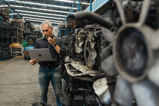 Supervisor with laptop inspecting used car engines in automotive spare parts warehouse