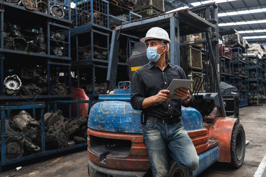 Warehouse manager in face mask using tablet while leaning on forklift in auto parts storage