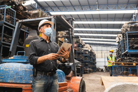 Warehouse manager in face mask using tablet while leaning on forklift in auto parts storage