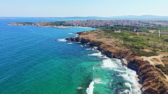 Aerial view shows the coastline with waves hitting the rocky shore. The clear blue water contrasts with the green landscape. A town is visible in the background under a bright sky.