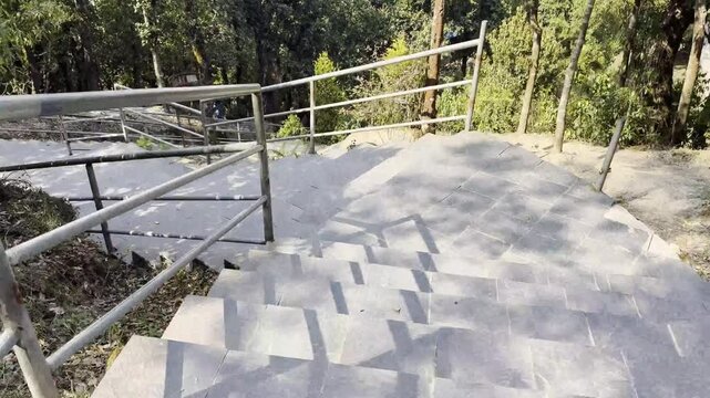 First person perspective walking down gray stone stairs with metal handrails in lush green forest park environment.