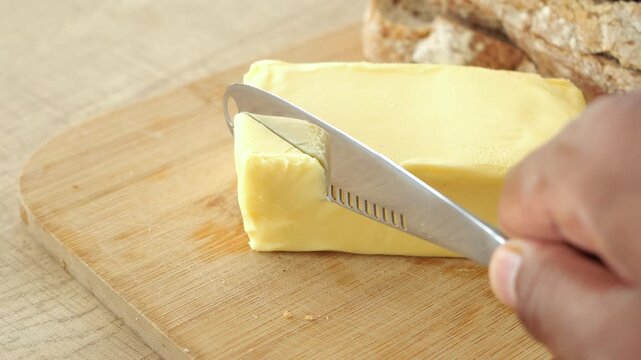 Person cutting piece of fresh yellow butter with steel knife on wooden board