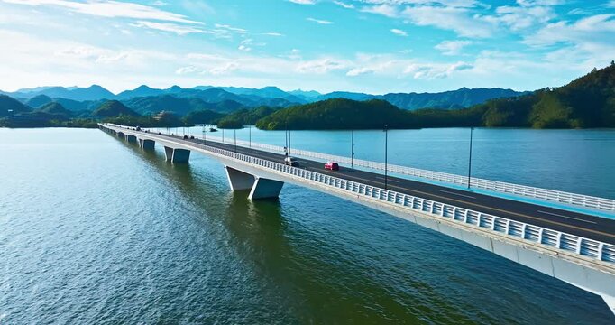 A long highway bridge spans across a wide lake under bright sunlight, with forested islands and distant hills visible beneath a blue cloudy sky.