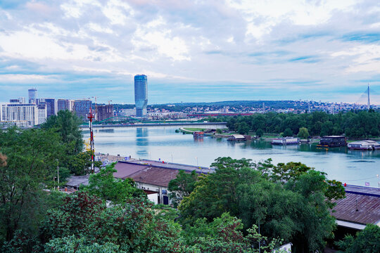 An elevated view over lush treetops frames the Sava River, moored floating barges, and the modern Belgrade Waterfront skyline featuring Kula Belgrade in Belgrade, Serbia