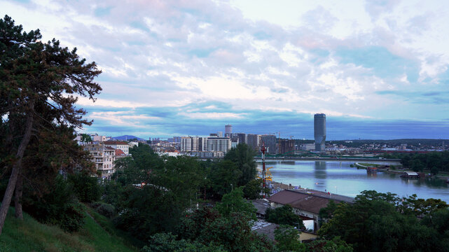 A high-angle view over lush green trees and the Sava River captures the Belgrade Waterfront development and Kula Belgrade skyscraper under an evening sky in Belgrade, Serbia