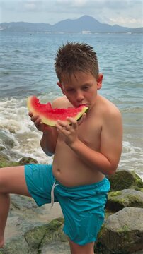 Happy blond Latino teenage boy eating a slice of red tropical watermelon on the golden beach of the Pacific coast in Manzanillo, Colima, Mexico vertical shot