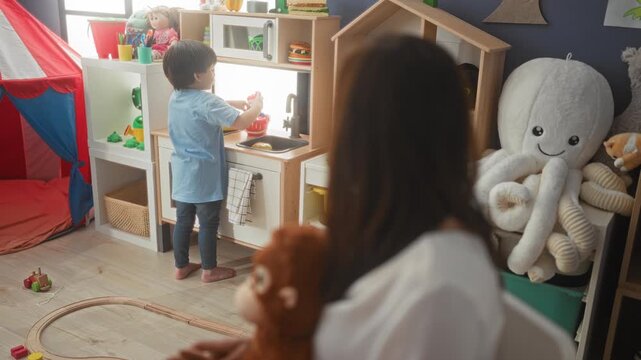 Woman holds stuffed monkey watching boy hold a green plate at a toy kitchen in a playroom; playful bonding.