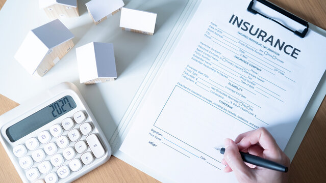 Hand signing an insurance form beside wooden house models and calculator, concept of home insurance, property protection, mortgage planning, financial security and real estate investment.