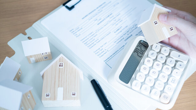Hand holding a wooden house above a calculator and contract documents, concept of home buying, mortgage calculation, property agreement, financial planning and real estate investment.