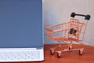 Miniature shopping cart next to a modern tablet with blank mockup screen on a wooden table, online shopping and e-commerce business concept with copy space. © Isman