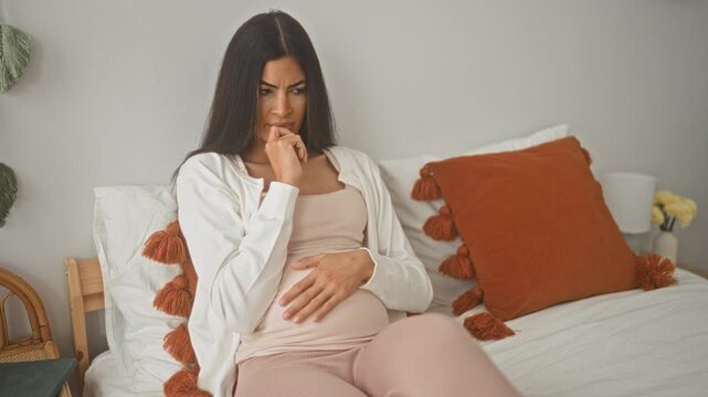 Woman with pregnant belly sitting on bed, hand resting on bump and finger to mouth thinking beside tasseled pillow in bedroom; maternal concern.