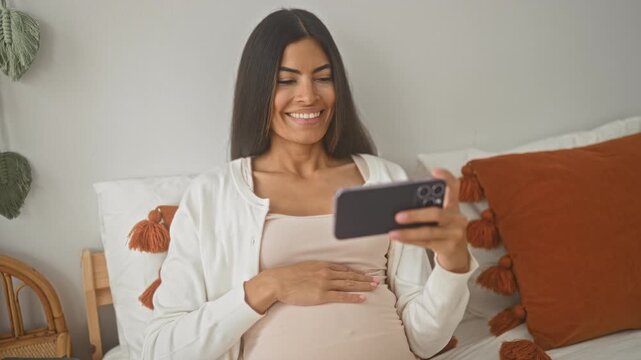 Pregnant woman sitting on bed, smiling, touching bare belly and holding smartphone, with tassel pillows and rattan chair visible inside building; maternal connection joyful.