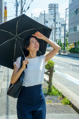 Japanese woman in her 30s holding a black parasol and looking up at the summer sky on a sunny street © Takeda Planning