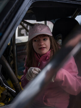 Young girl wearing a pink jacket and cap while sitting inside a modified folkrace car with a visible roll cage and racing seat.