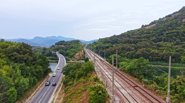 A road runs parallel to a railway track beside a river. Green trees cover the hills, and mountains are visible in the distance under a cloudy sky.