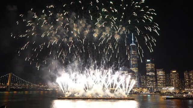 Emerald and sapphire fireworks illuminating the night sky over a glowing modern city skyline and river at midnight.