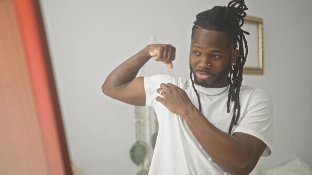 Young african american man flexes bicep, smooths white tee and inspects his reflection in a mirror in a minimalist studio bedroom setting; confidence.