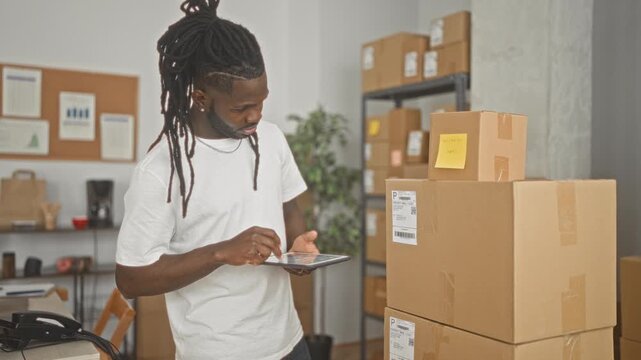 Man holding tablet and tapping screen beside stacked parcel boxes in a small business building while checking orders and labels; entrepreneurial confidence.