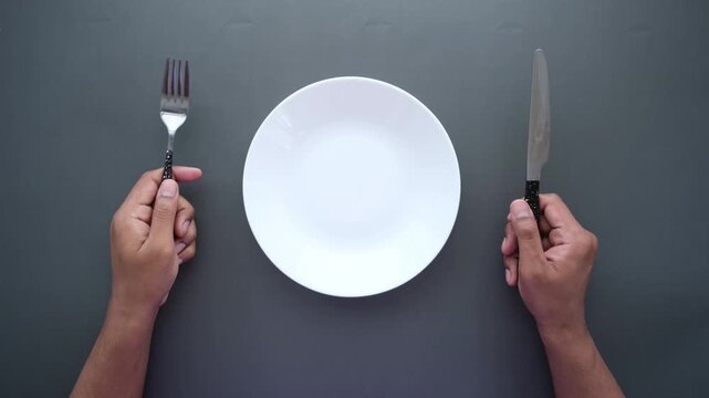 Hands holding silver fork and knife with empty white plate on dark gray background overhead view