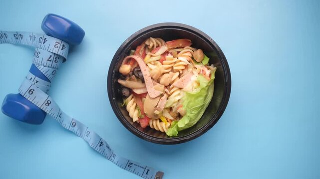 Top-down view of a colorful pasta salad bowl next to a dumbbell and measuring tape on a vibrant blue background, promoting fitness and healthy eating