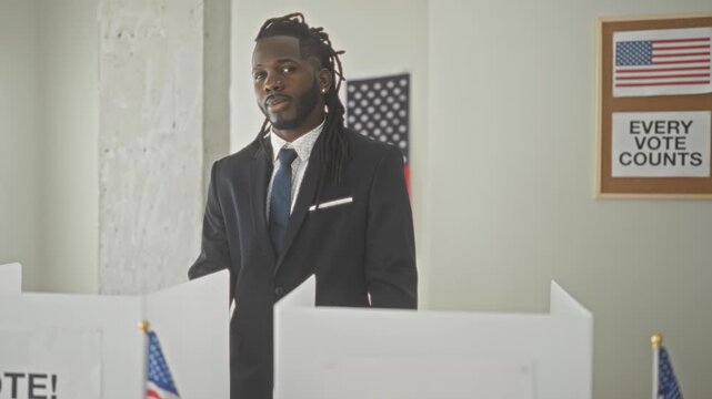 Man in suit standing at a voting booth beside american flag and voting partition in a polling building, smiling and looking toward camera; civic pride.