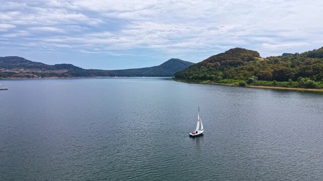 A sailboat glides across a lake with smooth water. Green hills are visible on both sides of the lake. The sky is covered with clouds. It is a quiet day for sailing.