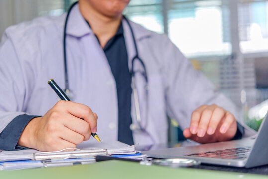 Healthcare professional writing clinical paperwork beside laptop with stethoscope for medical documentation administration and digital office workflow