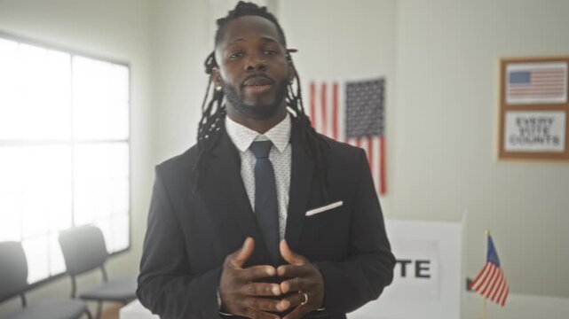 Man in suit speaking and gesturing with open hands toward ballot box and american flag in a voting building; voting civic pride.