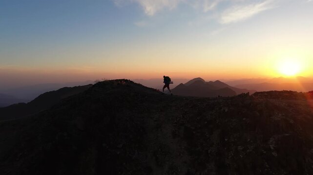 Alpinista con mochila de trekking ascendiendo por la cima de una monta&ntilde;a al atardecer con el sol detr&aacute;s, luz dorada intensa y una vista panor&aacute;mica &eacute;pica del horizonte