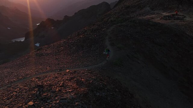 Alpinista con mochila de trekking ascendiendo por la cima de una monta&ntilde;a al atardecer con el sol detr&aacute;s, luz dorada intensa y una vista panor&aacute;mica &eacute;pica del horizonte