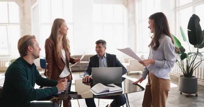 Diverse corporate colleagues gathering for informal briefing around office table collaborating on startup project reviewing documents and laptop sharing ideas laughing during relaxed team discussion