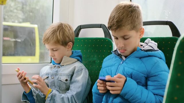 Two young boys sit side by side on green patterned public transport seats, both intently interacting with handheld smartphones, left boy in light denim jacket and blue hoodie holds yellow phone near