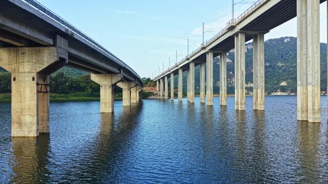 Two bridges span over a body of water with concrete pillars visible in the area. The scene shows clear skies and green landscape in the background.