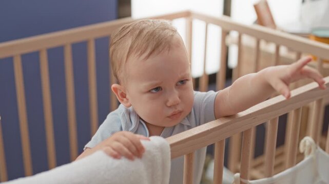 Blonde toddler boy reaches and points finger over wooden crib rail, hand holding towel and grasping railing in building; frustration longing.